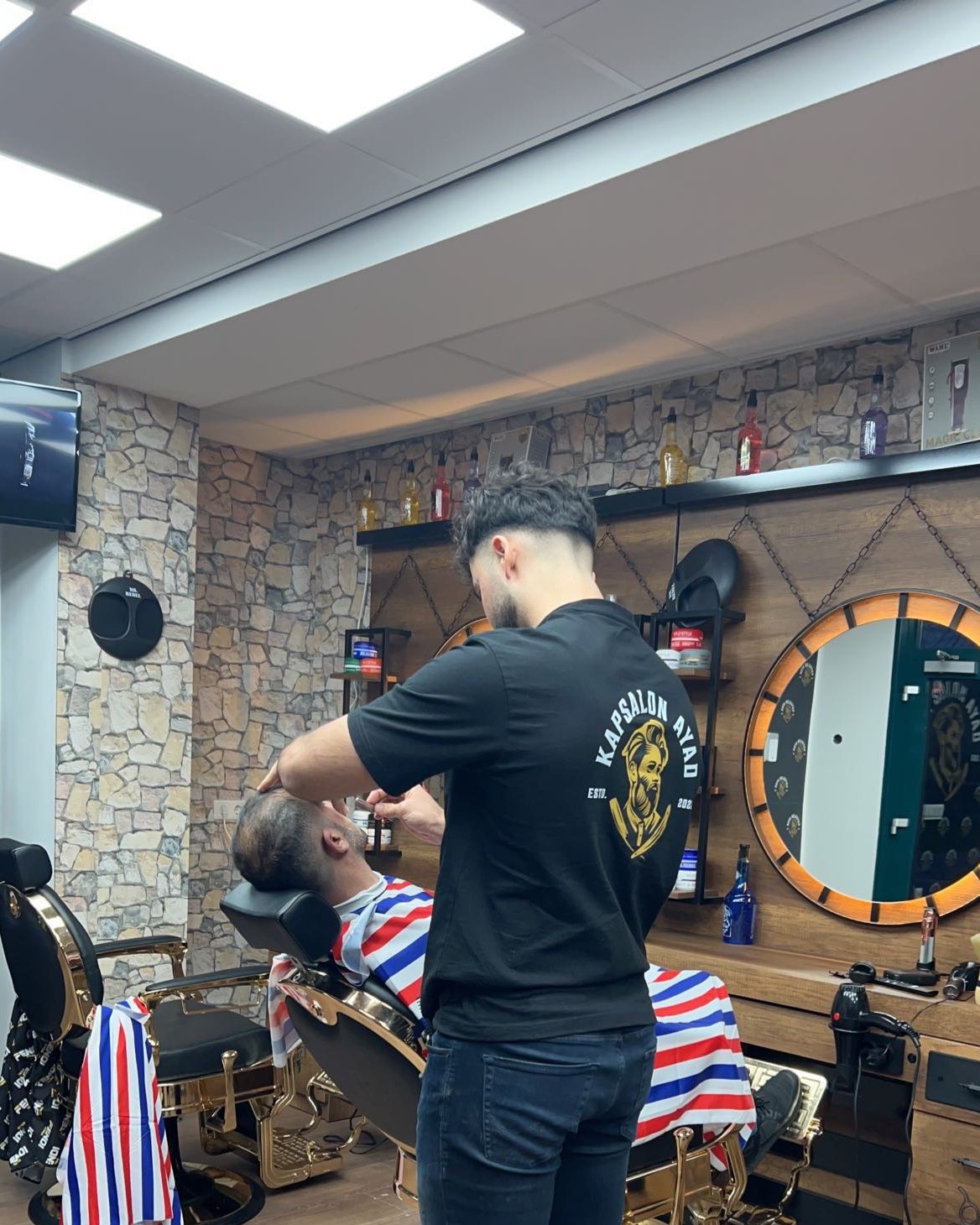 Een kapper in een zwart Kapsalon Ayad-shirt aan het werk bij een klant in de kappersstoel, naast een ronde spiegel met gouden rand