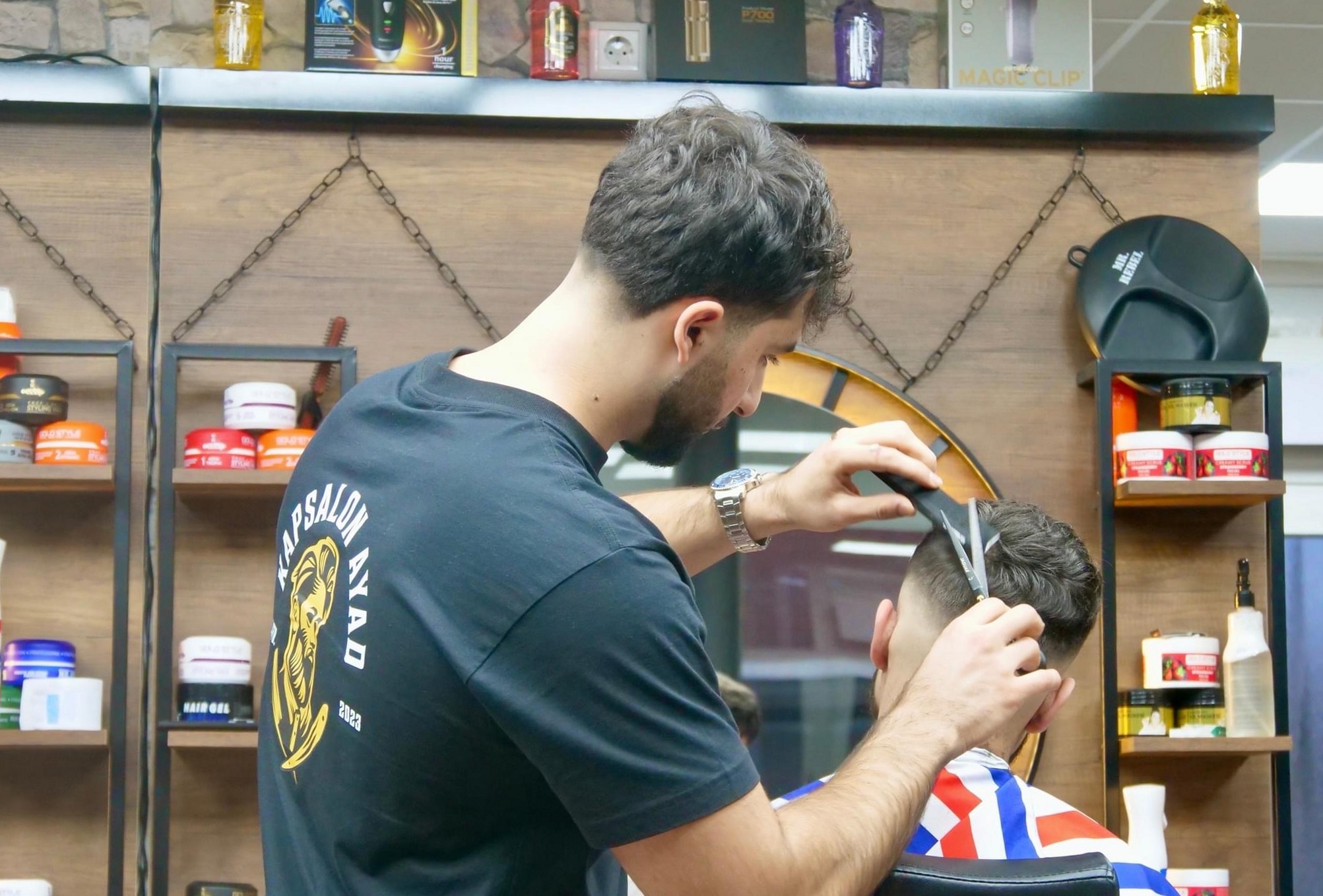 Een kapper in een zwart Kapsalon Ayad-shirt knipt een klant met de schaar, voor de wandkast met producten